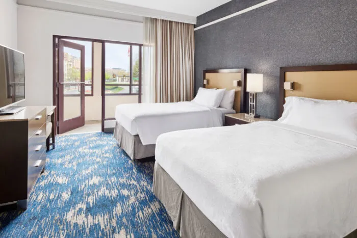 A hotel room with two neatly made double beds, a dark patterned accent wall, a TV on a dresser, large window doors with curtains, and blue patterned carpet. Natural light fills the room.