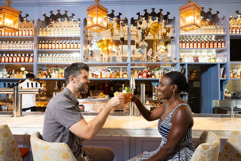 A man and woman sit at the marble bar insider the Founding Farmers Alexandria restaurant, smiling and toasting drinks.
