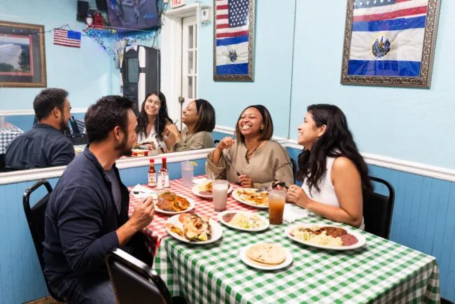 Four people sit at a restaurant table with Salvadoran food, smiling and laughing. The walls are decorated with Salvadoran flags, and colorful lights are visible in the background.