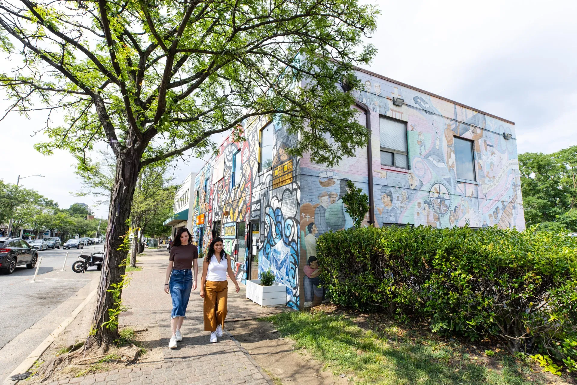 Two people walk along a sidewalk next to a building covered in colorful mural art, with trees, bushes, and parked cars lining the street on a bright day.