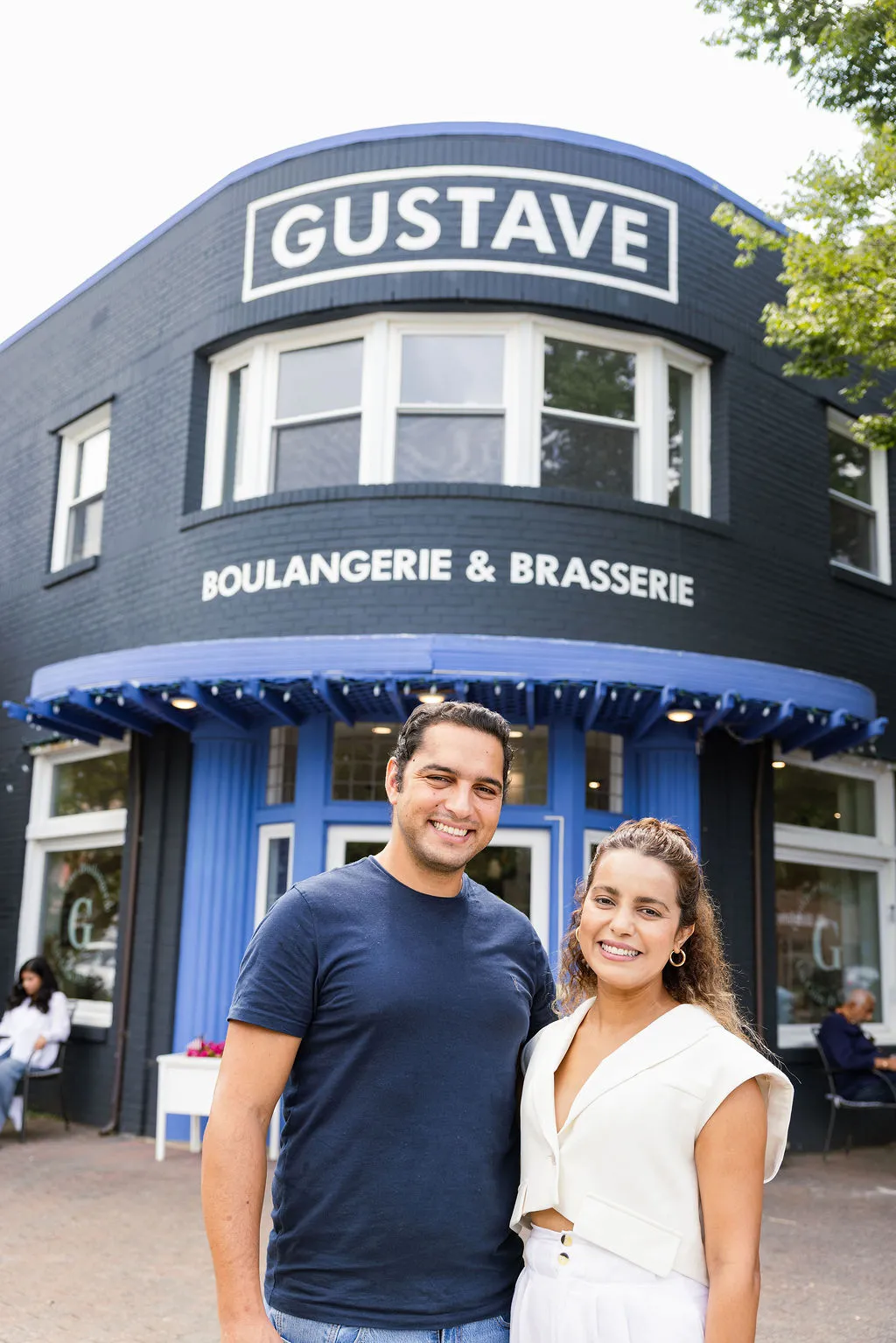 A man and woman smile in front of a navy blue building with a sign reading GUSTAVE and BOULANGERIE & BRASSERIE. People are seated outside near the entrance under a blue awning.