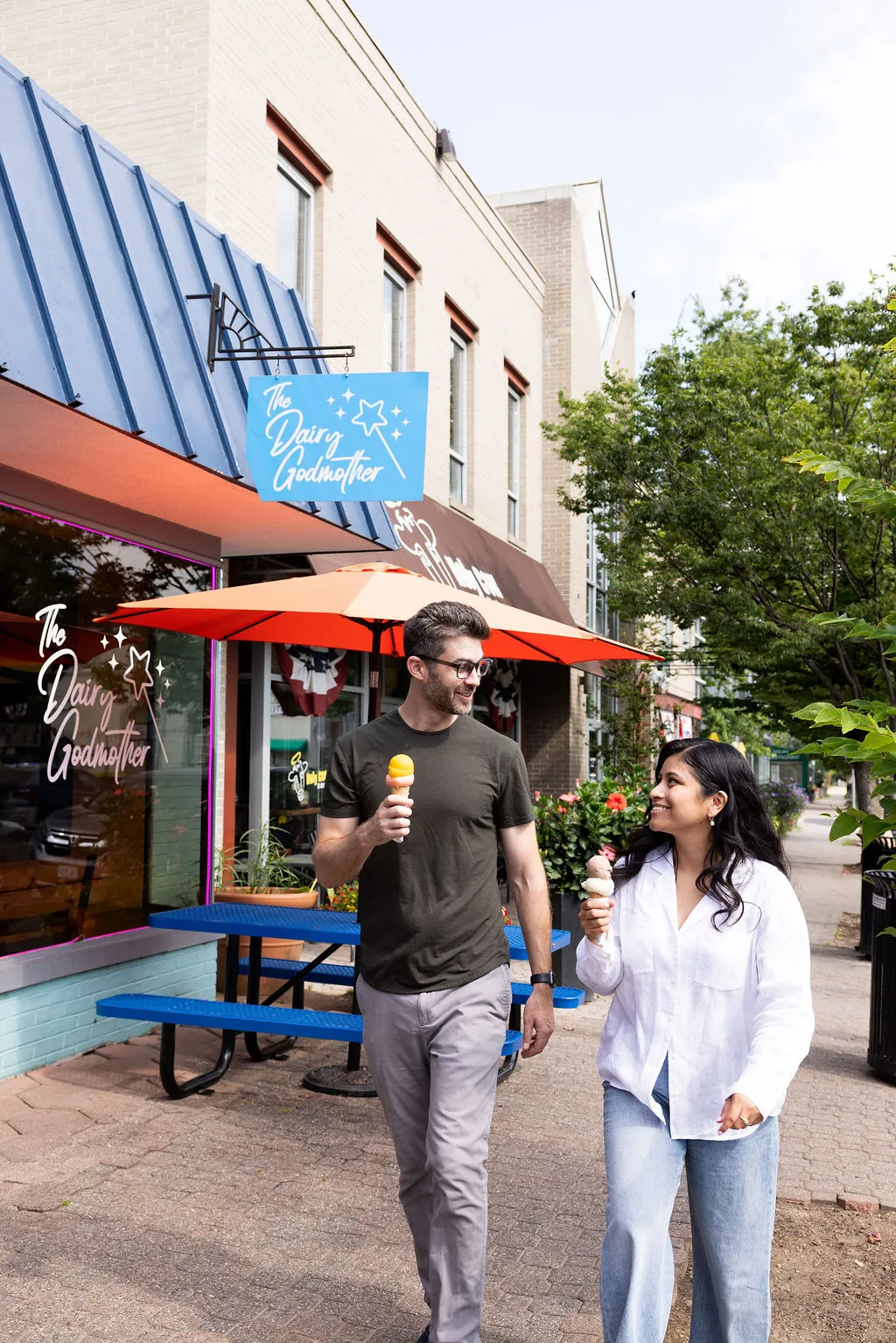 A man and woman smile as they walk outside an ice cream shop, each holding an ice cream cone. The shop has blue and orange decor, an outdoor seating area, and a sign that reads The Dairy Godmother.