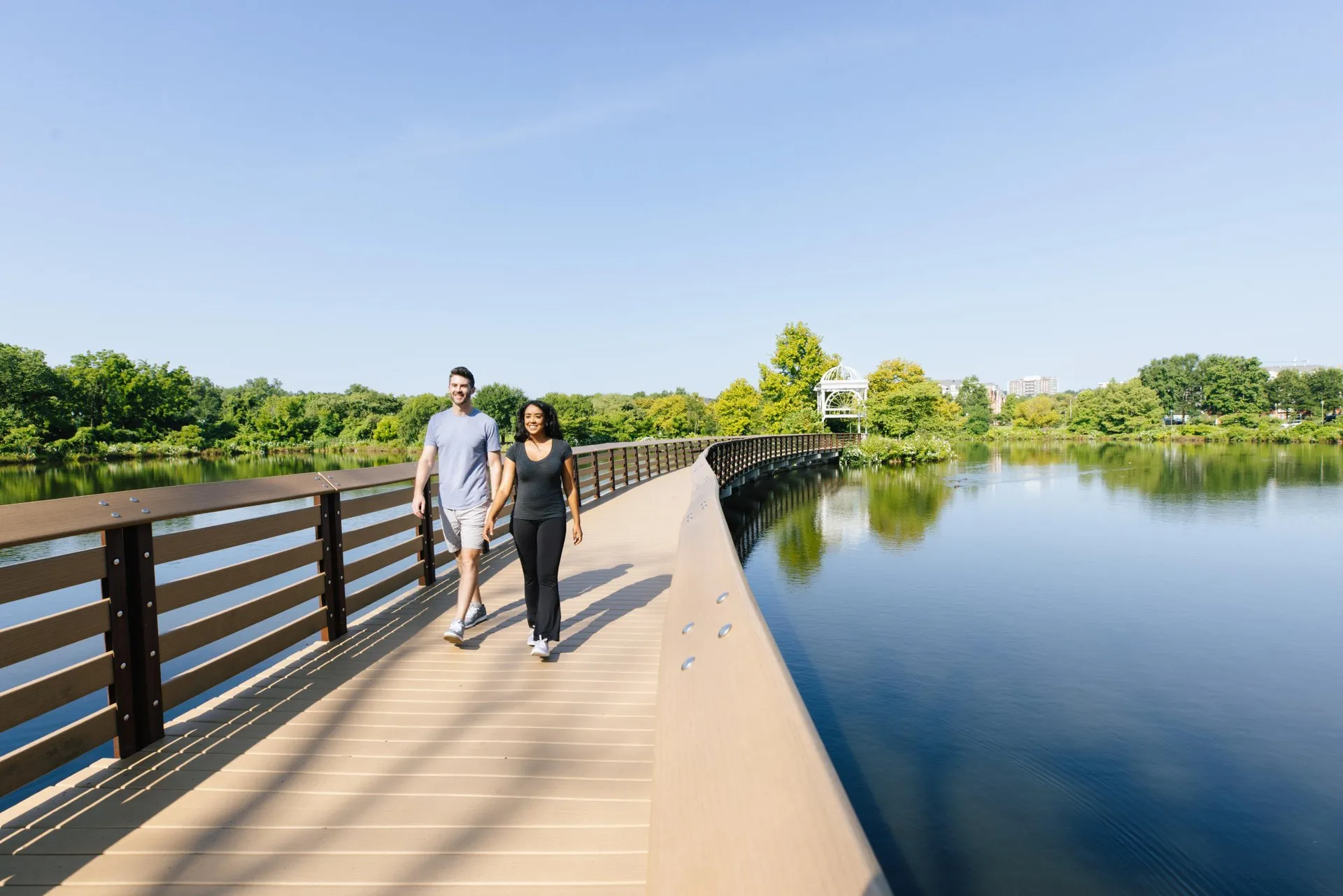 A man and woman walk together on a curved wooden bridge over a calm lake, surrounded by lush green trees and clear blue sky on a sunny day.