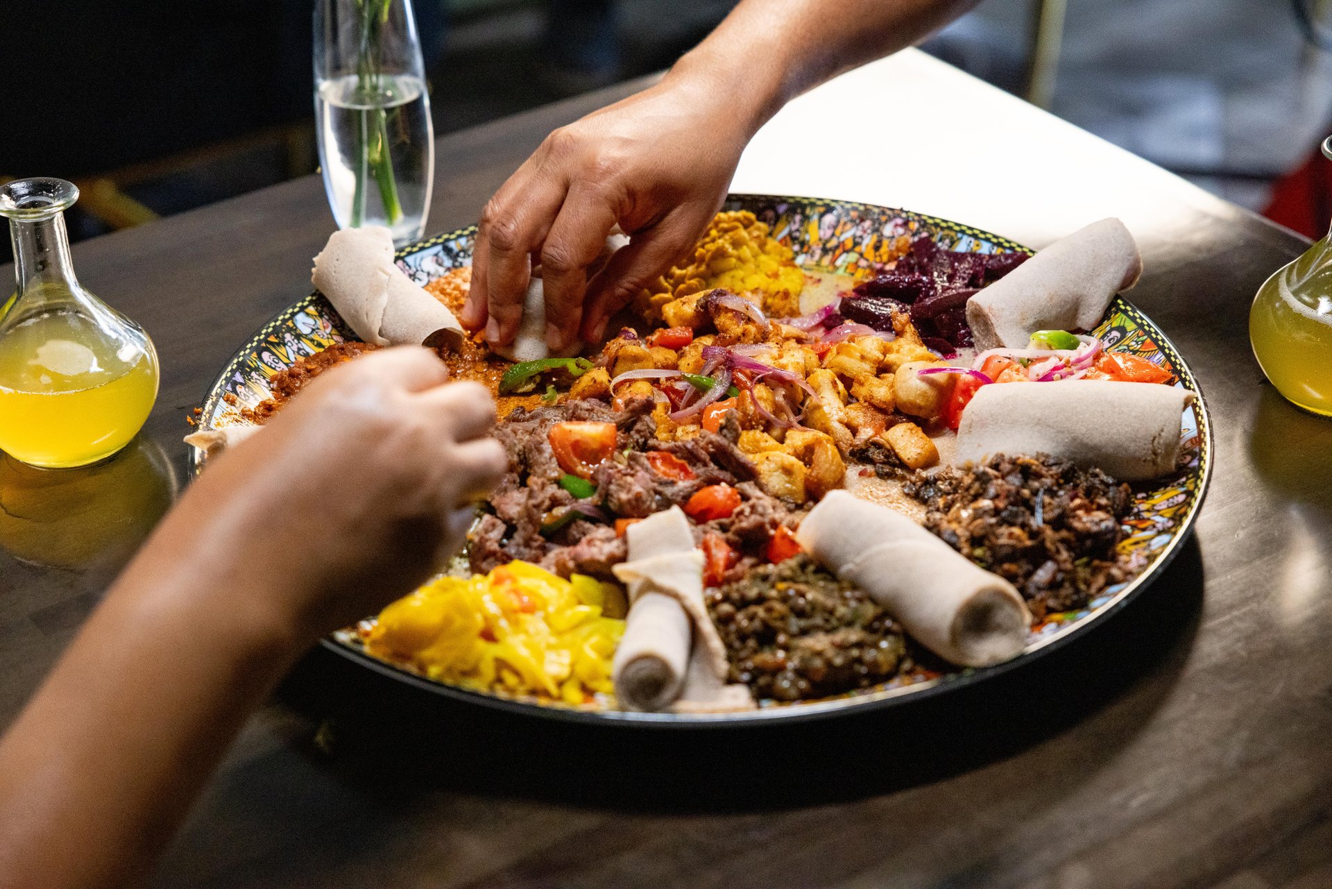 Two people use their hands to share a large platter of Ethiopian food, including injera, assorted stews, vegetables, and salad, on a dark table with drinks in the background.