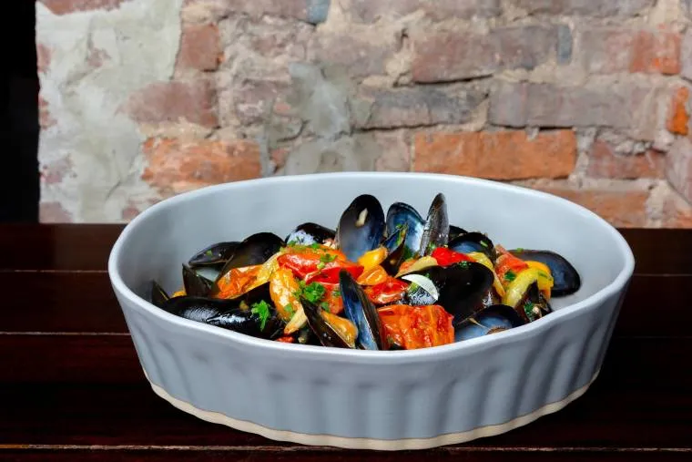 A white ceramic dish filled with cooked mussels, cherry tomatoes, and herbs sits on a dark wooden table, with a rustic brick wall in the background.