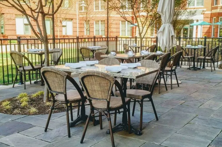 Outdoor patio with several tables and wicker chairs arranged neatly, each table set with plates and napkins. The area is paved with stone tiles and surrounded by a black metal fence and some greenery.