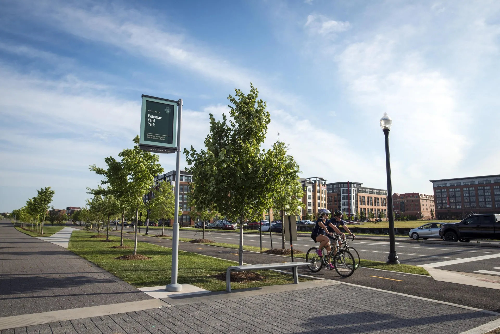 A person rides a bicycle on a paved path lined with young trees in the Potomac Yard neighborhood of Alexandria, VA.