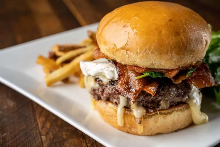 A close-up of a gourmet cheeseburger with melted cheese, crispy bacon, and lettuce on a brioche bun, served with French fries and a green salad on a white plate.