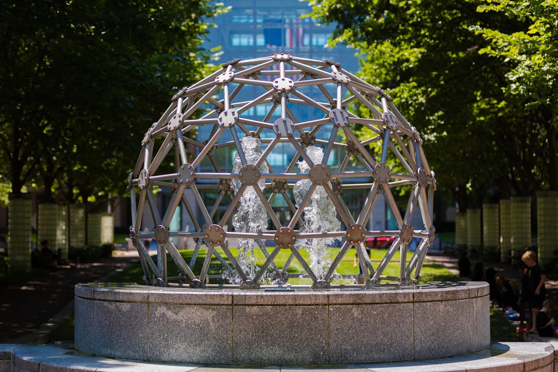 A metal geodesic dome sculpture stands over a circular stone fountain with water jets, surrounded by trees in an outdoor park setting.