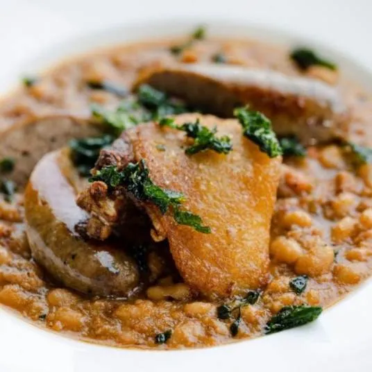 A close-up of a hearty bean stew topped with golden-brown crispy meat, browned sausages, and garnished with chopped herbs on a white plate.
