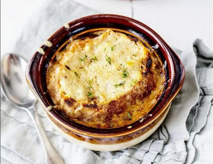 A bowl of French onion soup topped with a slice of toasted bread and melted, browned cheese, garnished with fresh thyme. A spoon and a light gray napkin are placed beside the bowl.