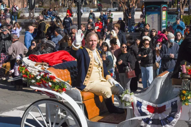 A man in historical attire rides in a decorated horse-drawn carriage, waving to a crowd. Spectators in winter clothing line the street, watching and taking photos. The scene appears festive, with patriotic bunting and flowers.