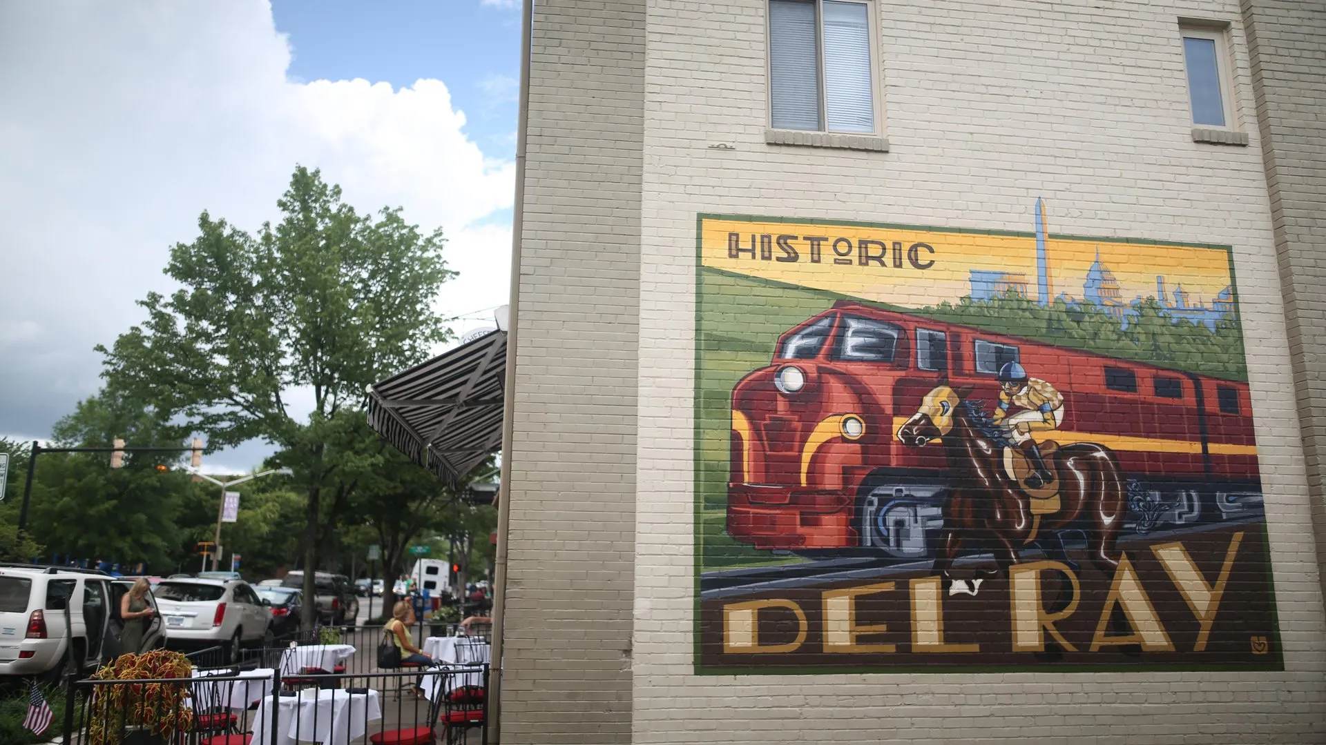A mural on a brick wall shows a red train and a rider on horseback with the words Historic Del Ray. To the left, a tree-lined street has parked cars and outdoor dining tables set up.