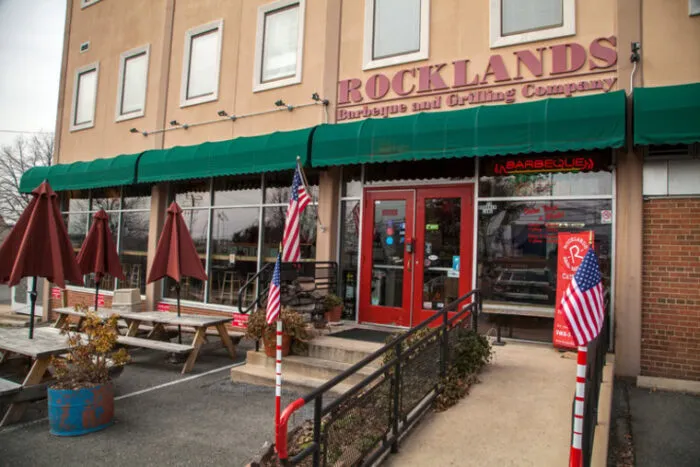 The exterior of Rocklands Barbeque and Grilling Company, featuring a green awning, red double doors, picnic tables, and American flags along the entrance ramp.