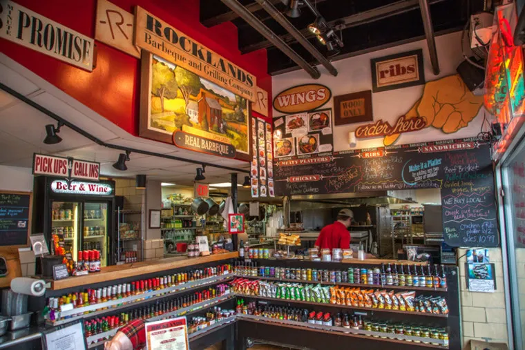 A colorful barbecue restaurant interior with sauce bottles on shelves, chalkboard menus, and wall signs advertising ribs, wings, and barbecue. A staff member stands behind the counter near a kitchen area.