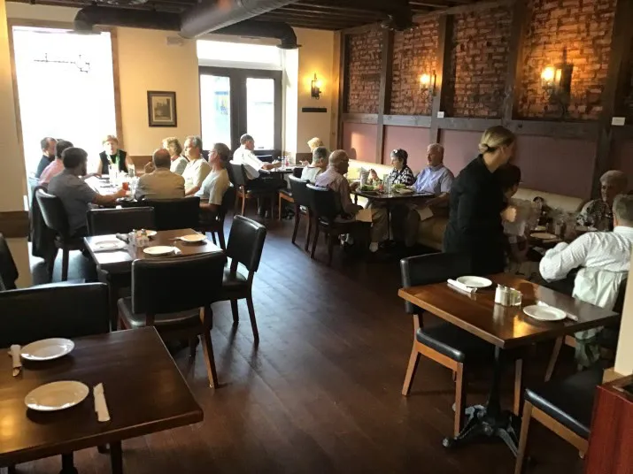 A cozy restaurant with people dining at tables, engaged in conversation. A server stands near a table, and the room features wooden floors, dark chairs, exposed brick walls, and warm lighting.