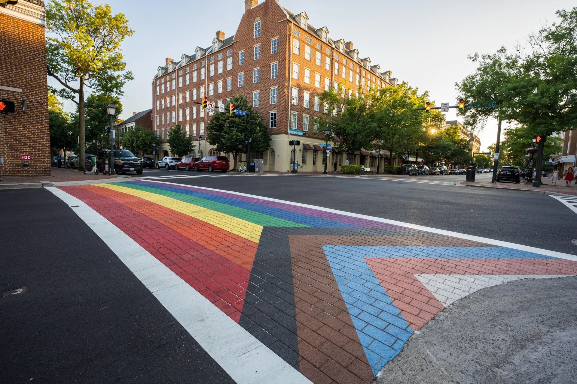 A crosswalk painted with the Progress Pride flag colors spans a brick street in an urban area, with a large red-brick building and trees in the background.