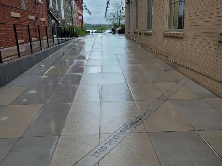A wet, empty walkway lined with brick buildings on both sides. String lights hang overhead, and “1789 HISTORIC HIGHLINE” is engraved on the pavement. Trees and a river are visible in the distance.