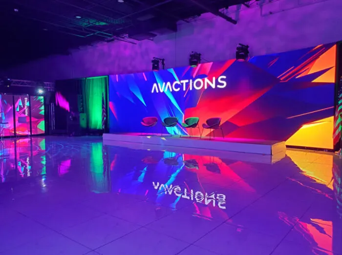 A brightly lit stage with three modern chairs and a colorful, abstract LED screen displaying ACTIONS in bold text, reflecting vibrant colors onto a glossy floor in an indoor event space.