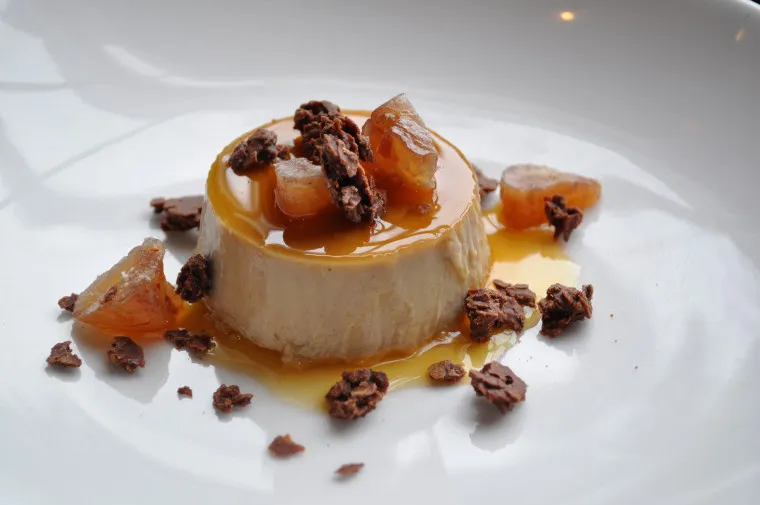 A round flan topped with caramel sauce, garnished with candied fruit pieces and chocolate crumbles, served on a white plate.