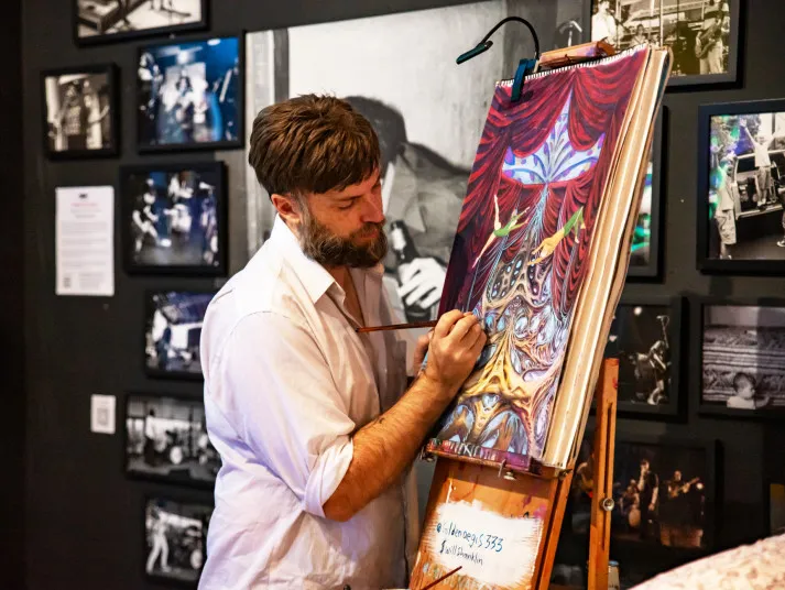 A bearded man with a paintbrush focused on painting a colorful canvas on an easel inside an art gallery.