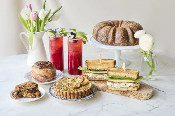 Assorted spring brunch spread with sandwiches, quiche, bundt cake, and tulips on a marble table.