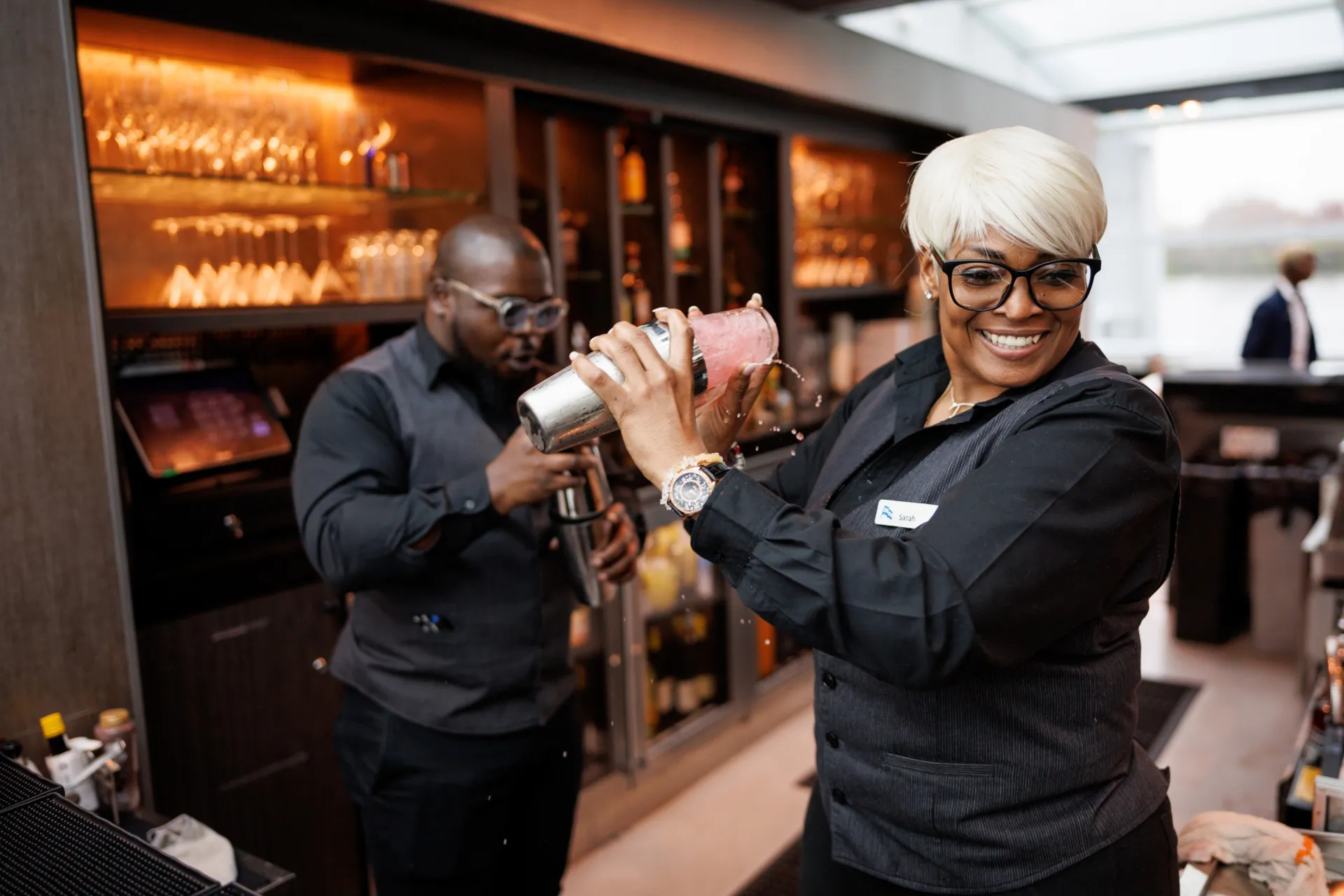 A smiling bartender with white hair and glasses preparing a pink cocktail in a shaker on a City Cruises ship.