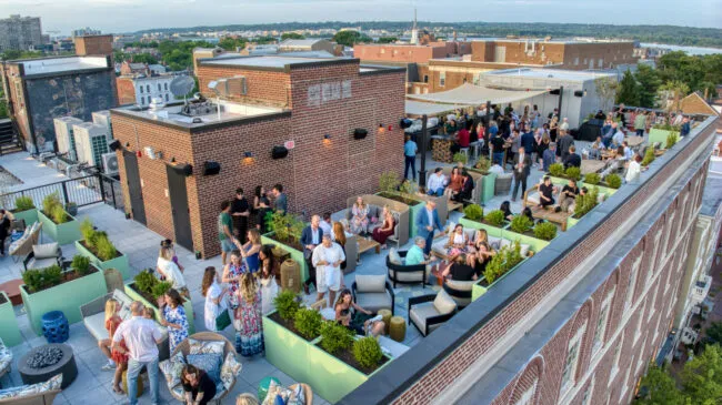 Crowded rooftop lounge at Hotel Heron with people sitting in green planters and outdoor seating.