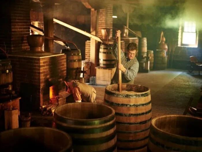 Men in historical attire working at a whiskey distillery with large wooden barrels and a brick hearth.
