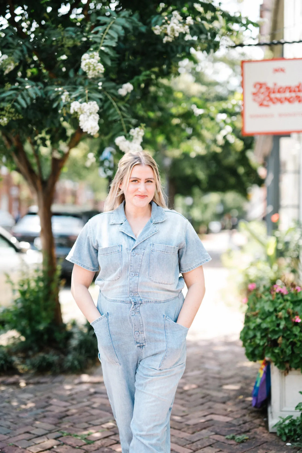 A woman smiles with her hands in her pockets on a sunny, tree-lined sidewalk in front of a sign that reads, 'Friends to Lovers'