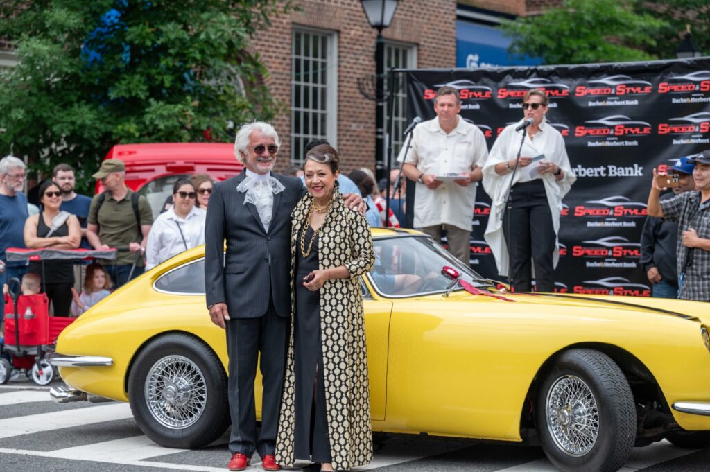A couple posing next to a yellow vintage sports car at the Festival of Speed & Style.