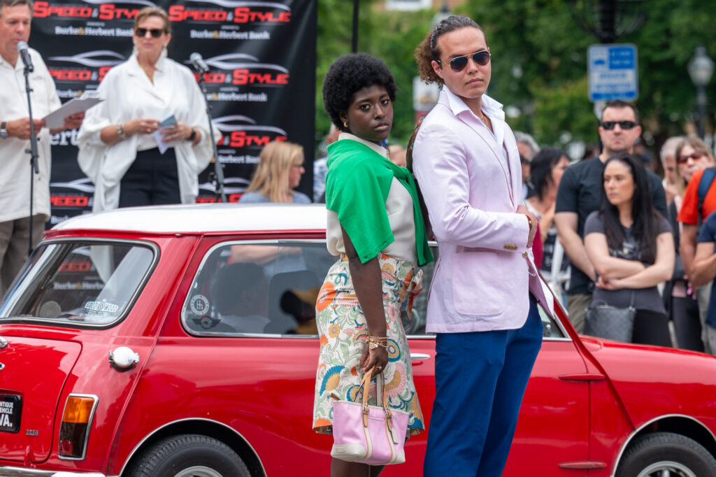 Fashionably dressed couple posing in front of a red vintage car at the Festival of Speed & Style.