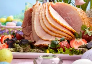Slices of glazed ham served on a platter with strawberries, asparagus, and colorful Easter eggs.