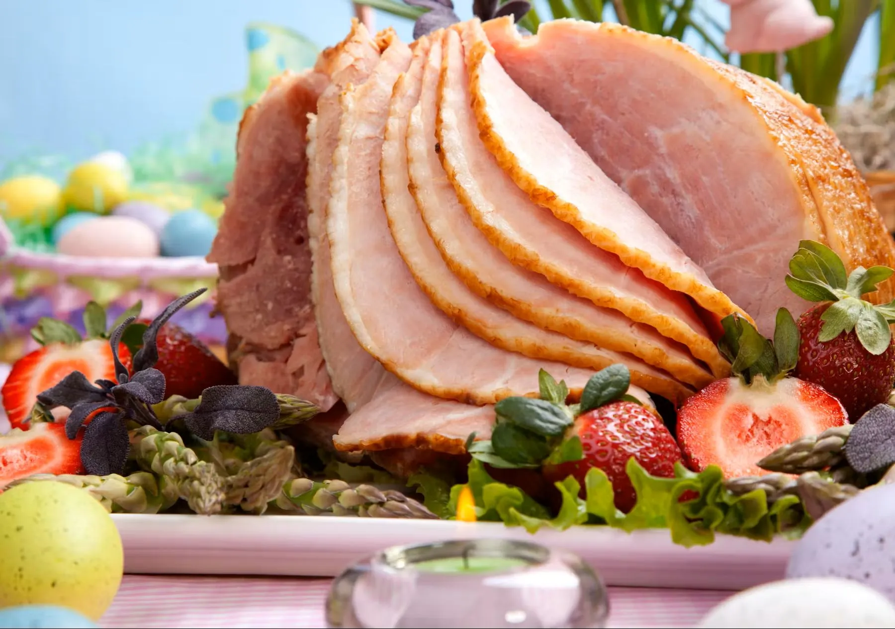 Slices of glazed ham served on a platter with strawberries, asparagus, and colorful Easter eggs.