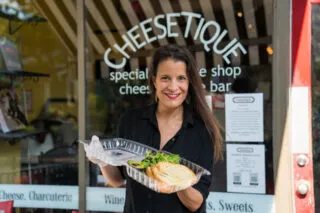 Women with long dark hair stands in front of the Cheesetique storefront holding a plastic take-out container of bread and cheese.