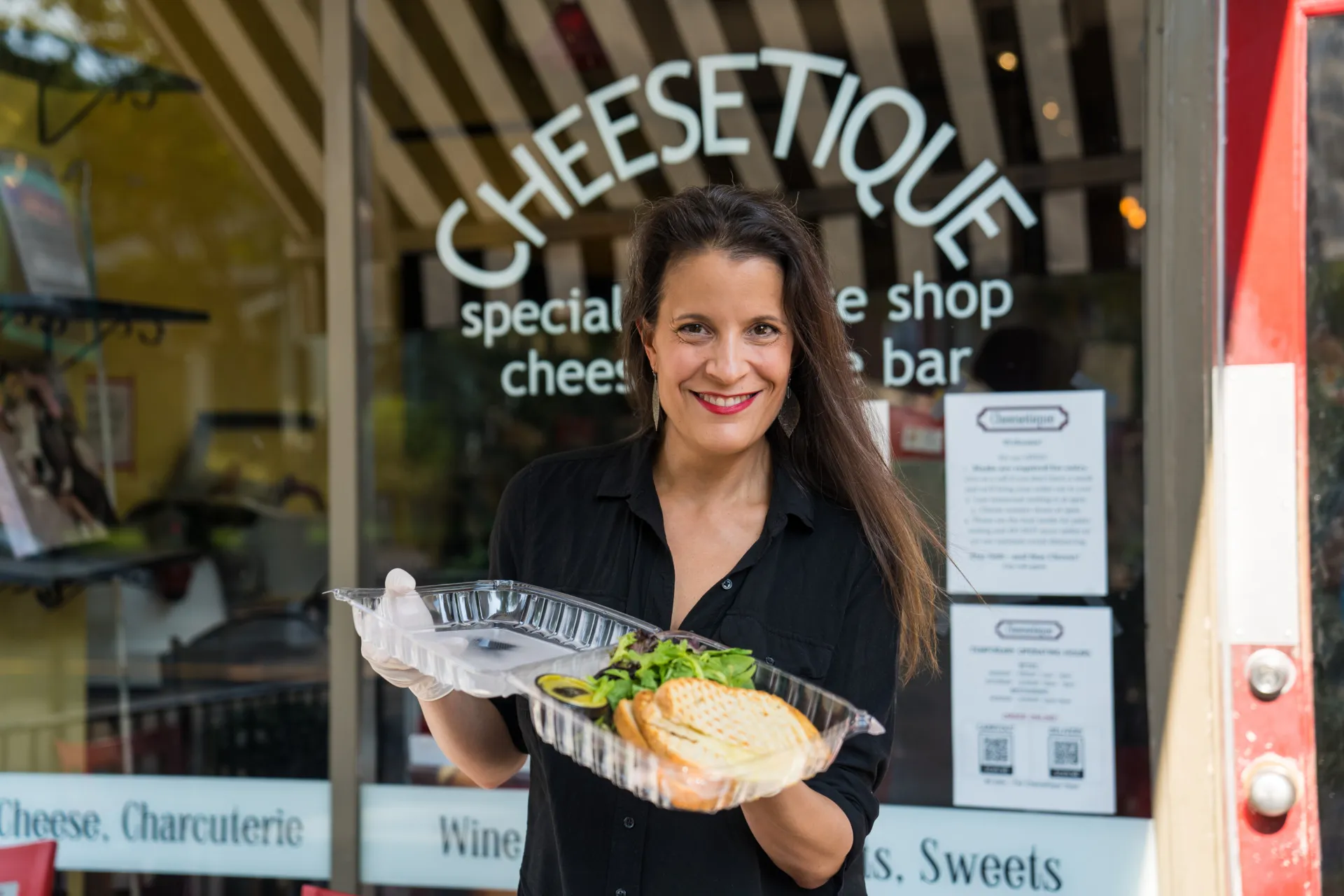 Women with long dark hair stands in front of the Cheesetique storefront holding a plastic take-out container of bread and cheese.