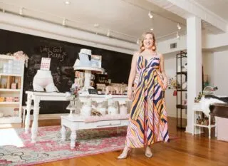 A blonde woman in a colorful, patterned maxi dress smiling inside Ms. Moxie's Moon Shop.