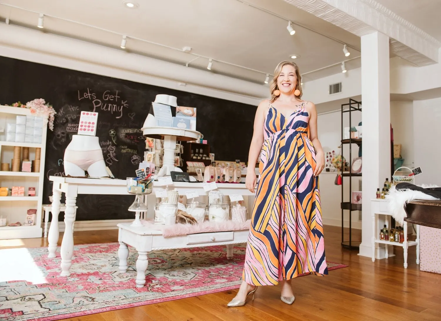 A blonde woman in a colorful, patterned maxi dress smiling inside Ms. Moxie's Moon Shop.