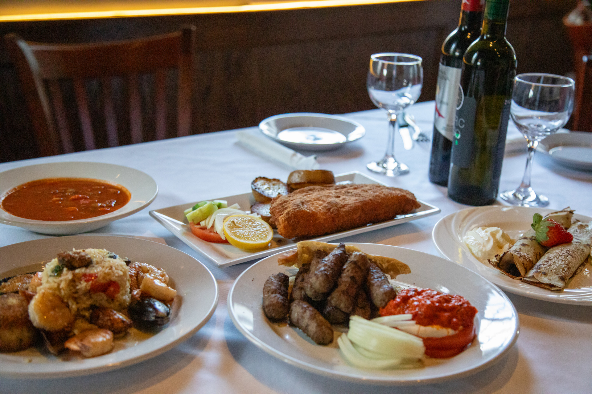 Several plates of food and a couple of wine bottles sit on a table adorned with white linens.