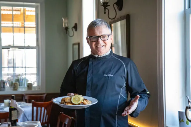 A man in a black chef's shirt holds a plate of food at the Old House Cosmopolitan Grill
