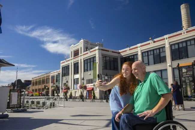 A young woman in a blue dress stands next to a man in a green shirt using a wheelchair to take a selfie in front of the Torpedo Factory at the Alexandria waterfront on a sunny day.
