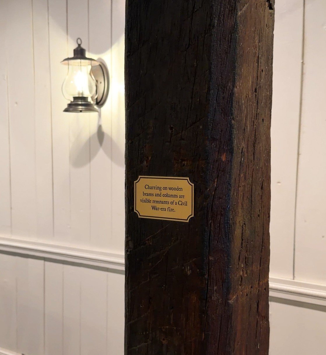 A charred wooden beam in front of a white paneled wall denotes that it is a remnant from a Civil War-era fire. A lantern mounted to the wall is illuminated in the background.