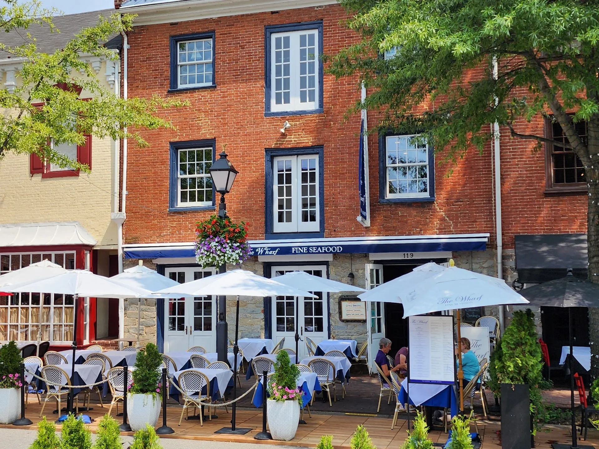A sunny shot of the exterior of The Wharf Fine Seafood Restaurant on Historic King Street, featuring outdoor dining tables with white umbrellas and crisp white linens, plus flower planters.
