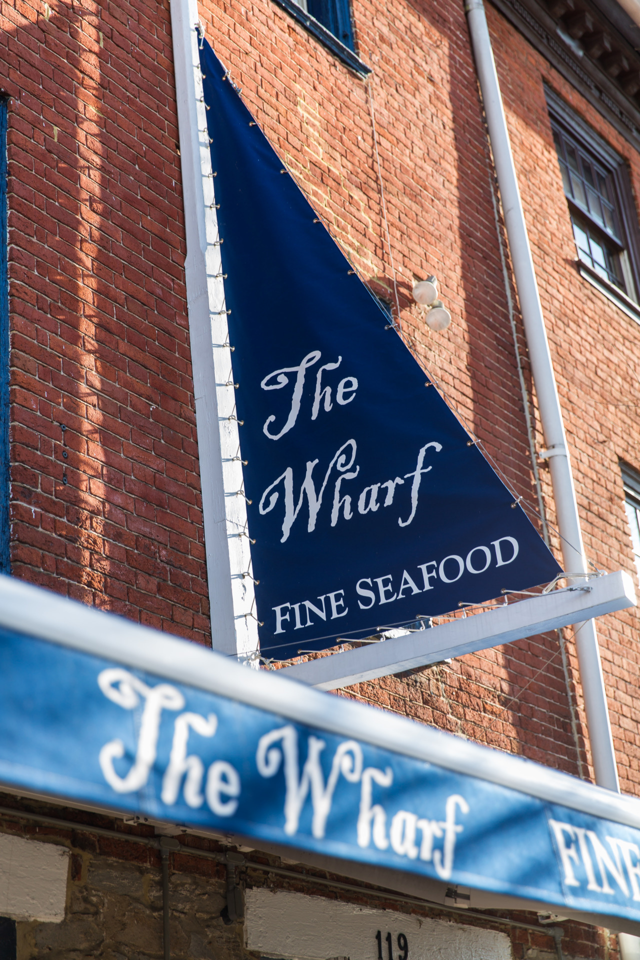 An exterior sign for The Wharf Fine Seafood is modeled after a sailboat, with navy sails and white lettering in front of the building's brick facade.