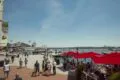 People walking along the paved waterfront promenade next to outdoor restaurant dining with red umbrellas at the Alexandria marina.