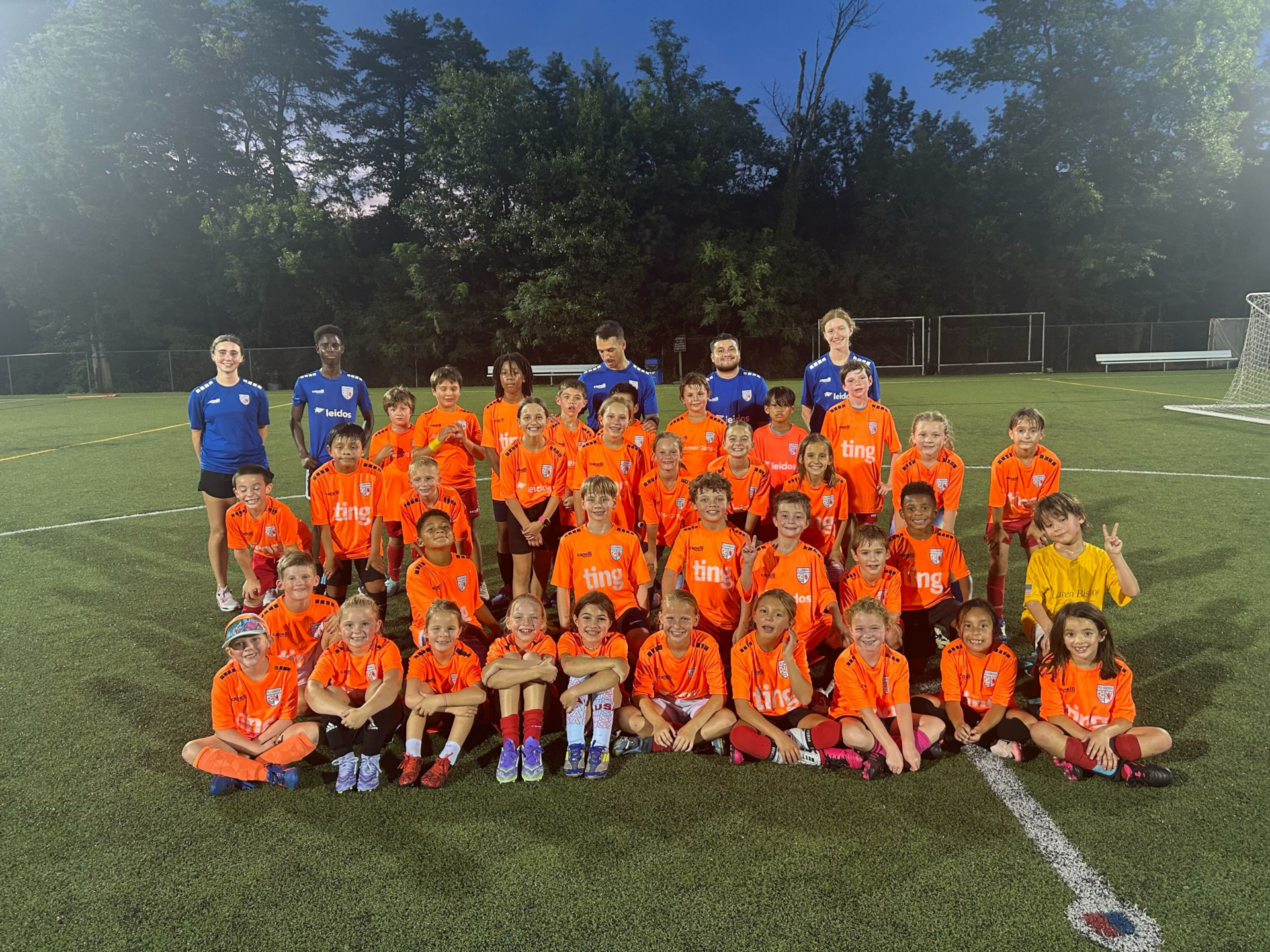 A group photo of young children in orange soccer uniforms sitting and standing together on a green field with their coaches at dusk.