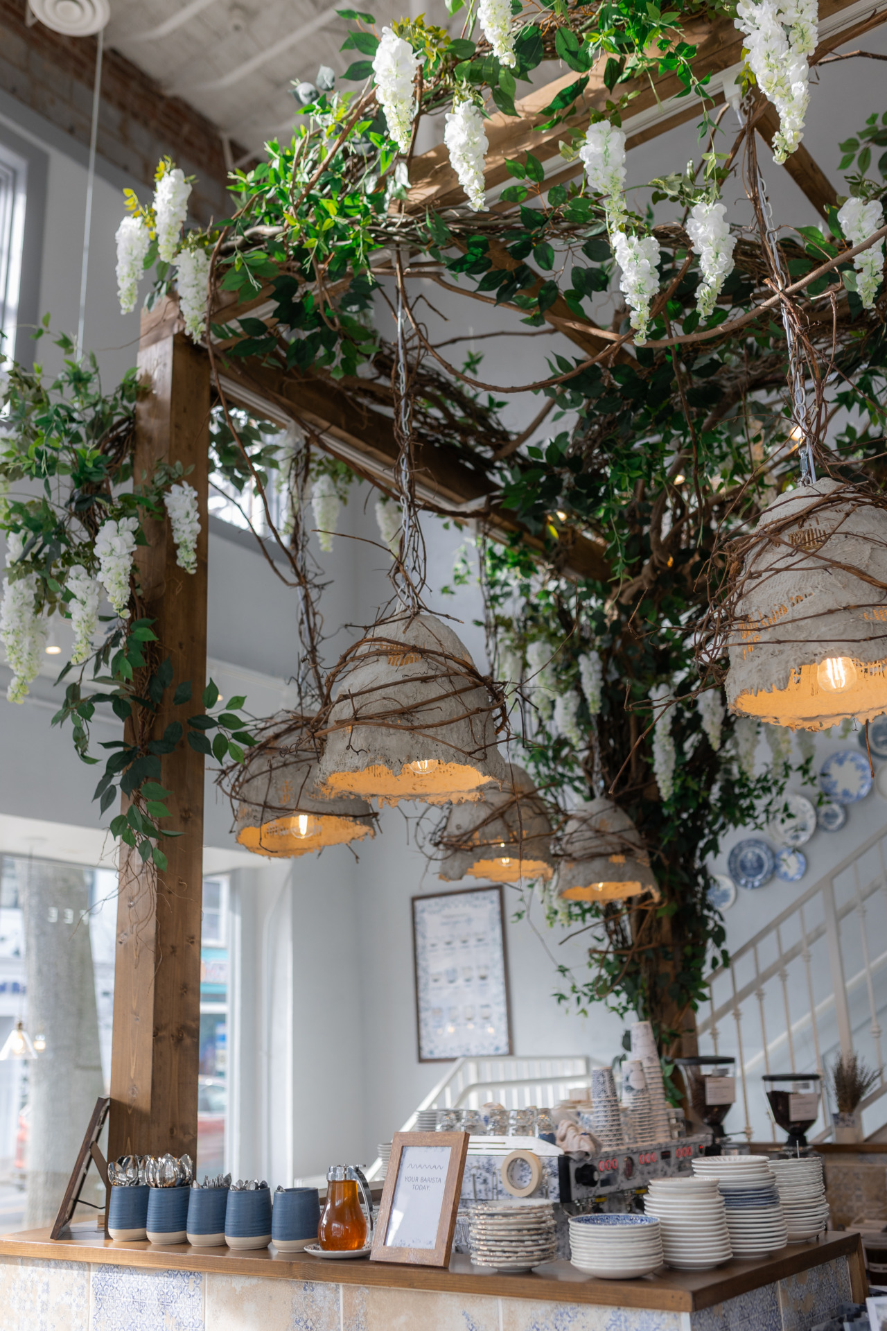 Woven basket light fixtures hanging under lush floral and green foliage arches inside Maman café.