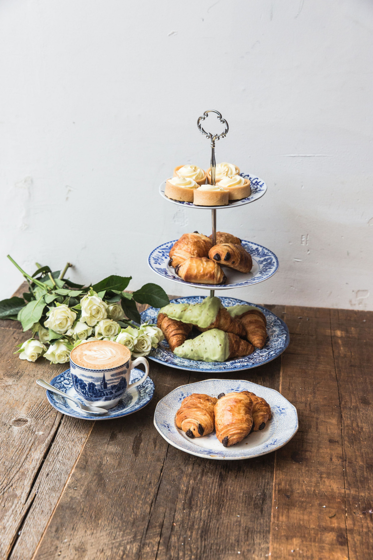 A three-tiered serving stand filled with assorted pastries and croissants next to a latte and white flowers on a wooden table.