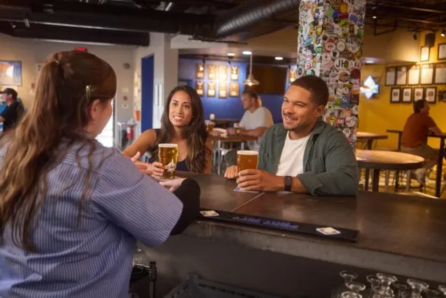 A smiling man and woman sit at a bar counter with beers, talking to a female bartender.