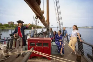 An 18th-century costumed guide gesturing and speaking to passengers gathered on the deck of the Tall Ship Providence.